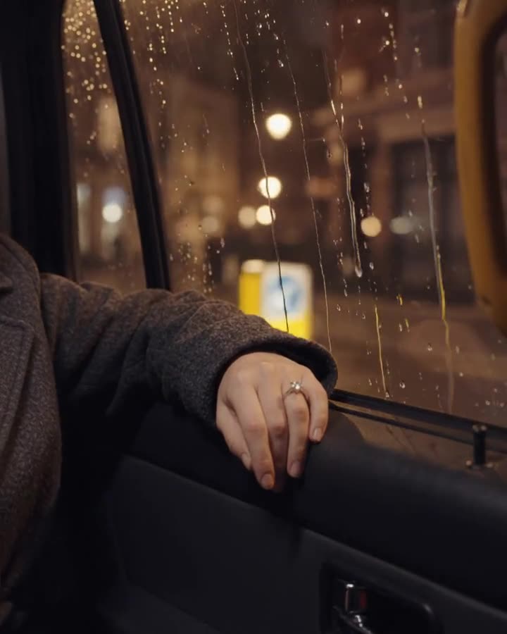 A hand wearing a solitaire rests by a rain-streaked taxi window at night.